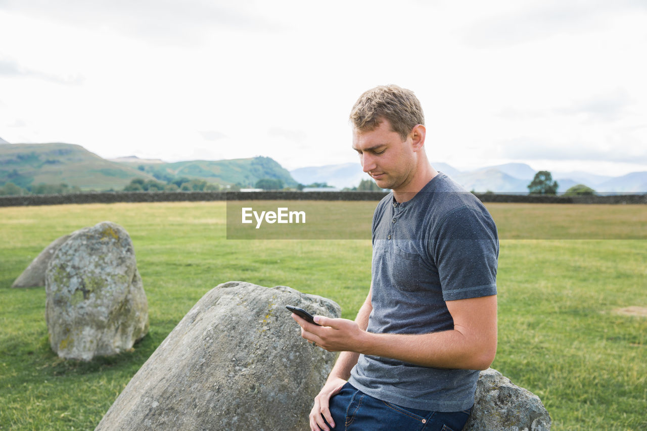 Young man using mobile phone while standing by rock on field