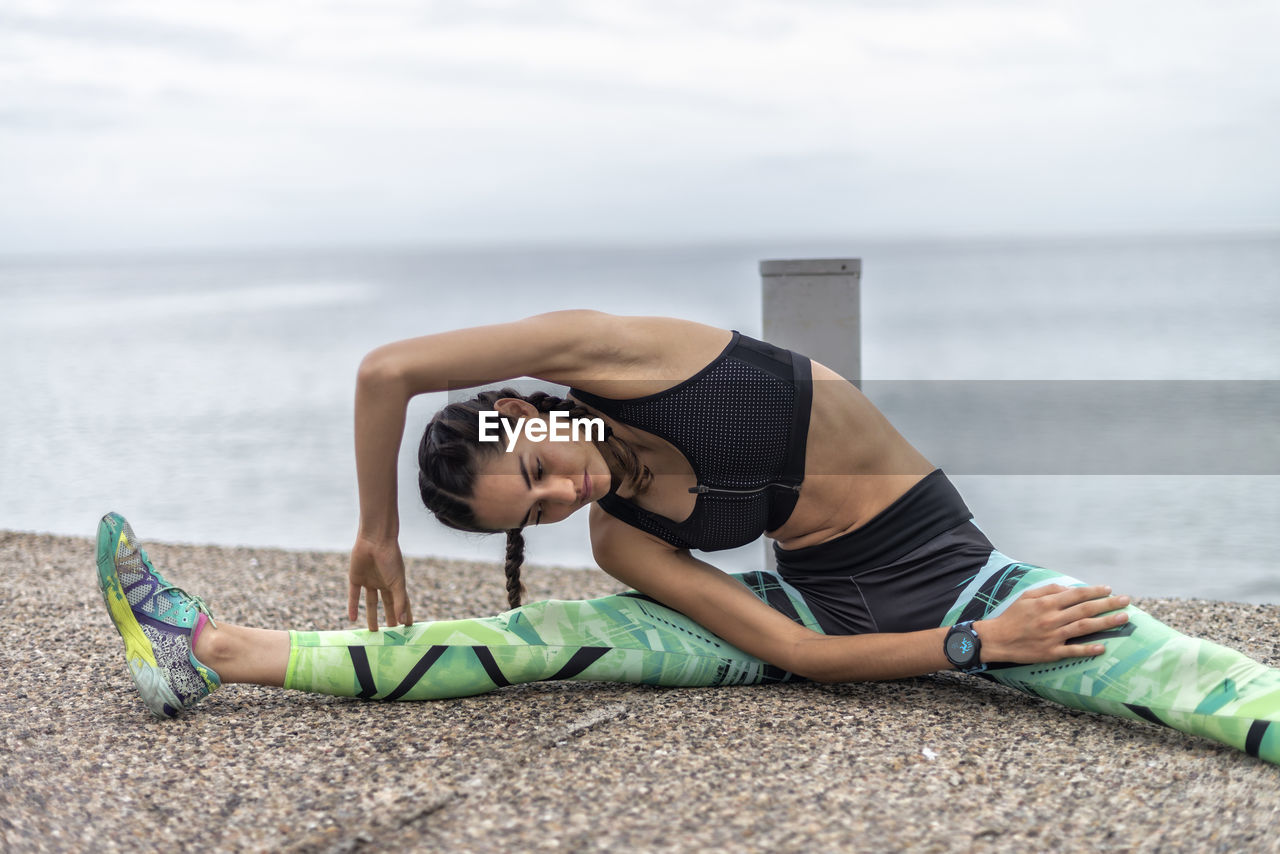 Fit female athlete sitting on embankment and doing side bend while stretching muscles and warming up during training