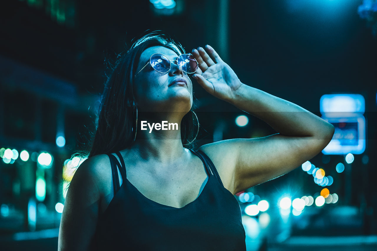 PORTRAIT OF YOUNG WOMAN STANDING AGAINST ILLUMINATED LIGHT