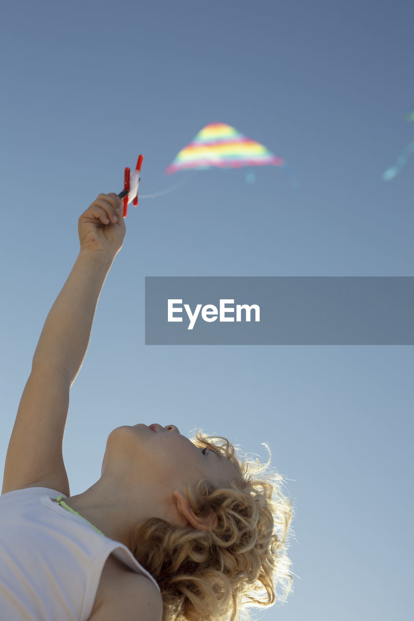 From below unrecognizable cute little boy with curly blond hair launching colorful kite under cloudless blue sky on sunny summer day