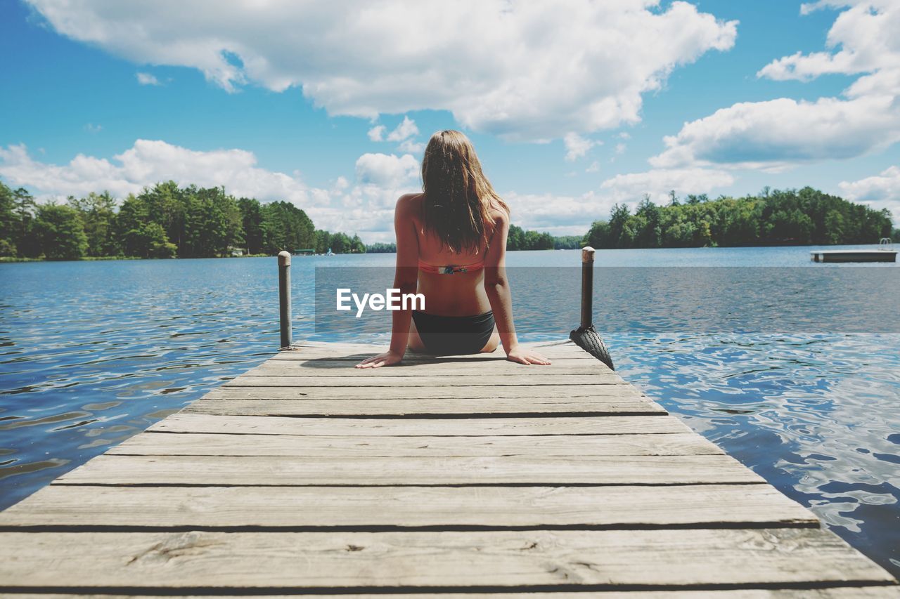 Rear view of teenage girl sitting on pier over lake against sky