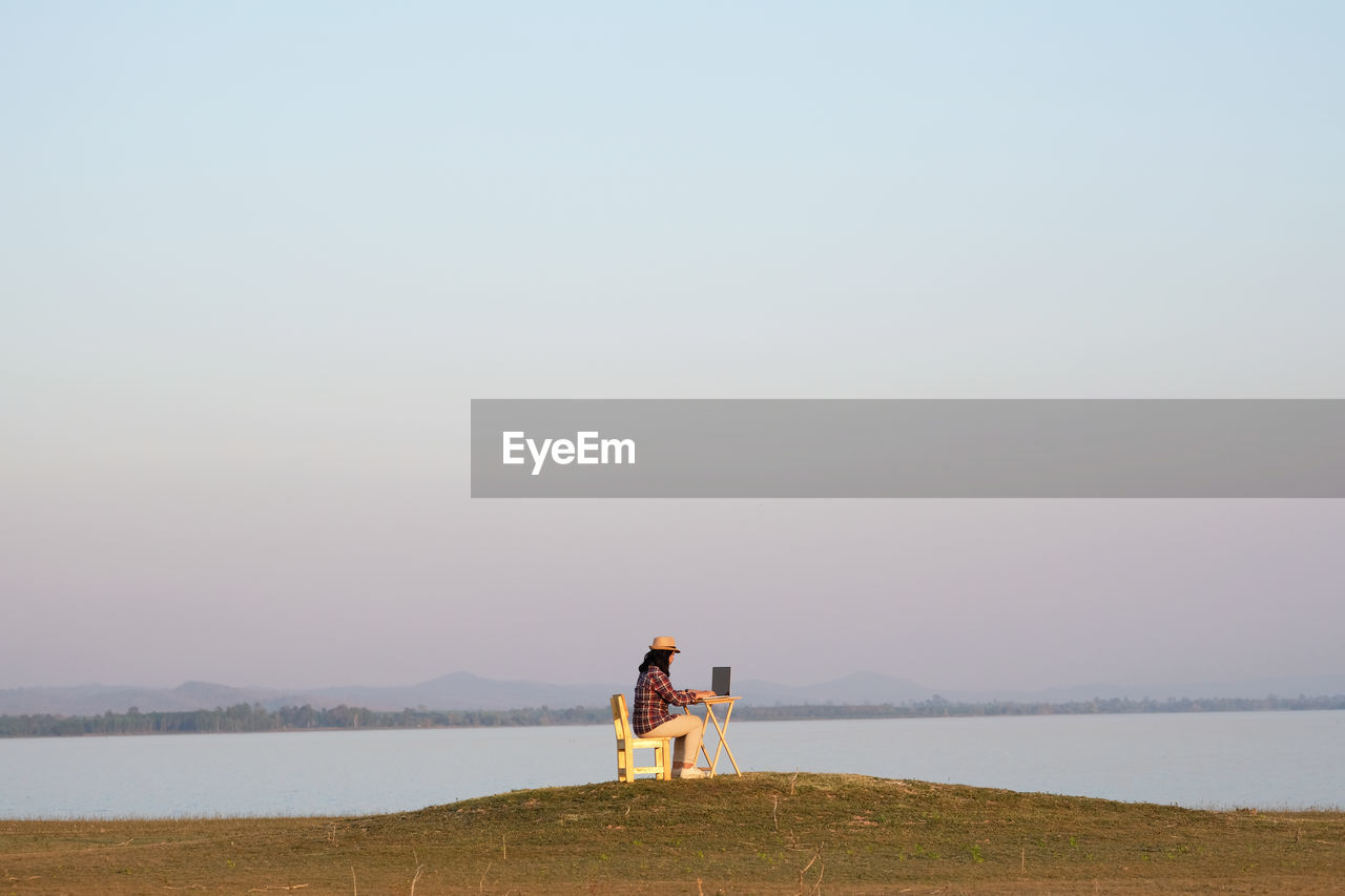 Woman using laptop over table sitting on grass by lake against clear sky