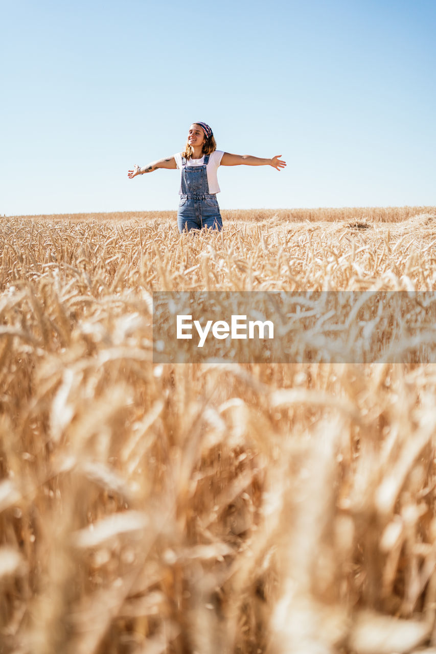 Delighted female in denim overalls standing in wheat field with outstretched arms and enjoying nature