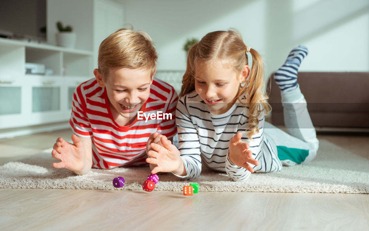 Cheerful siblings playing with dice while lying on carpet at home