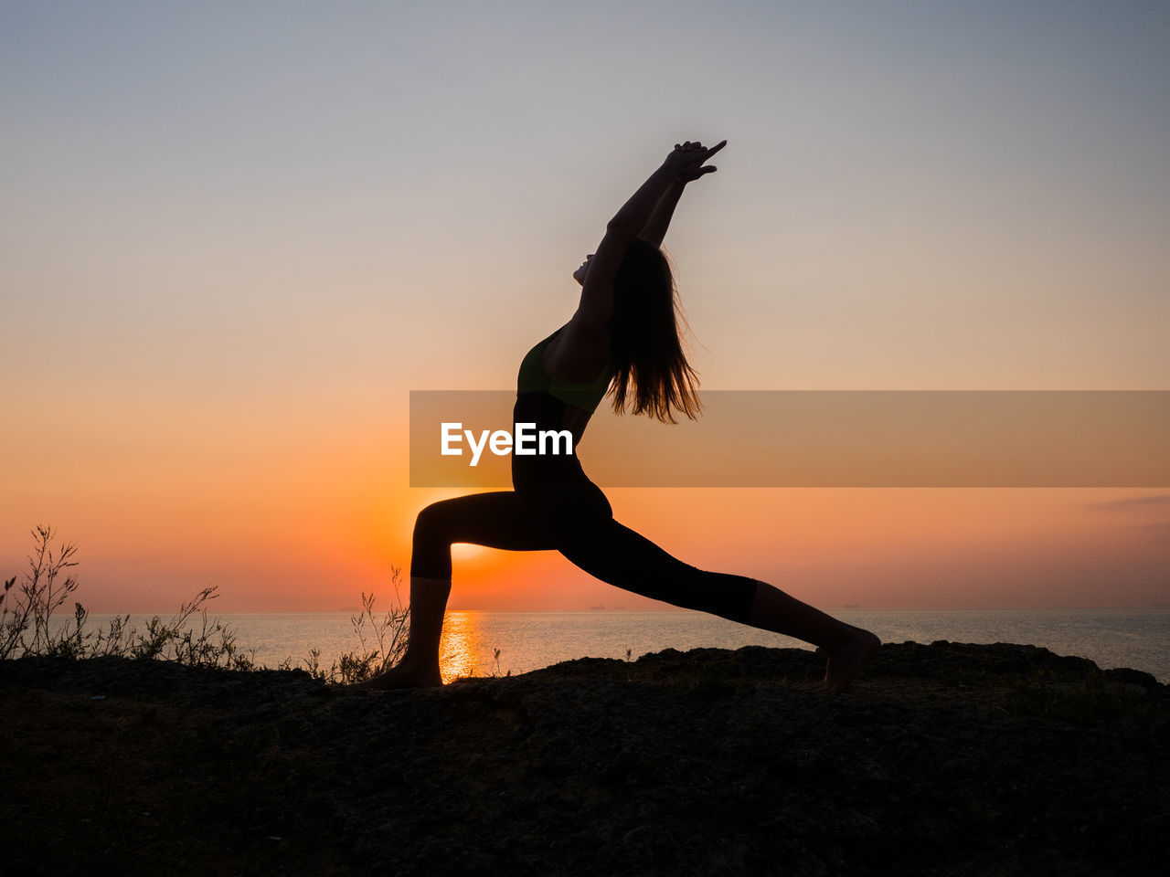 Side view of woman doing exercising on field against sea during sunset