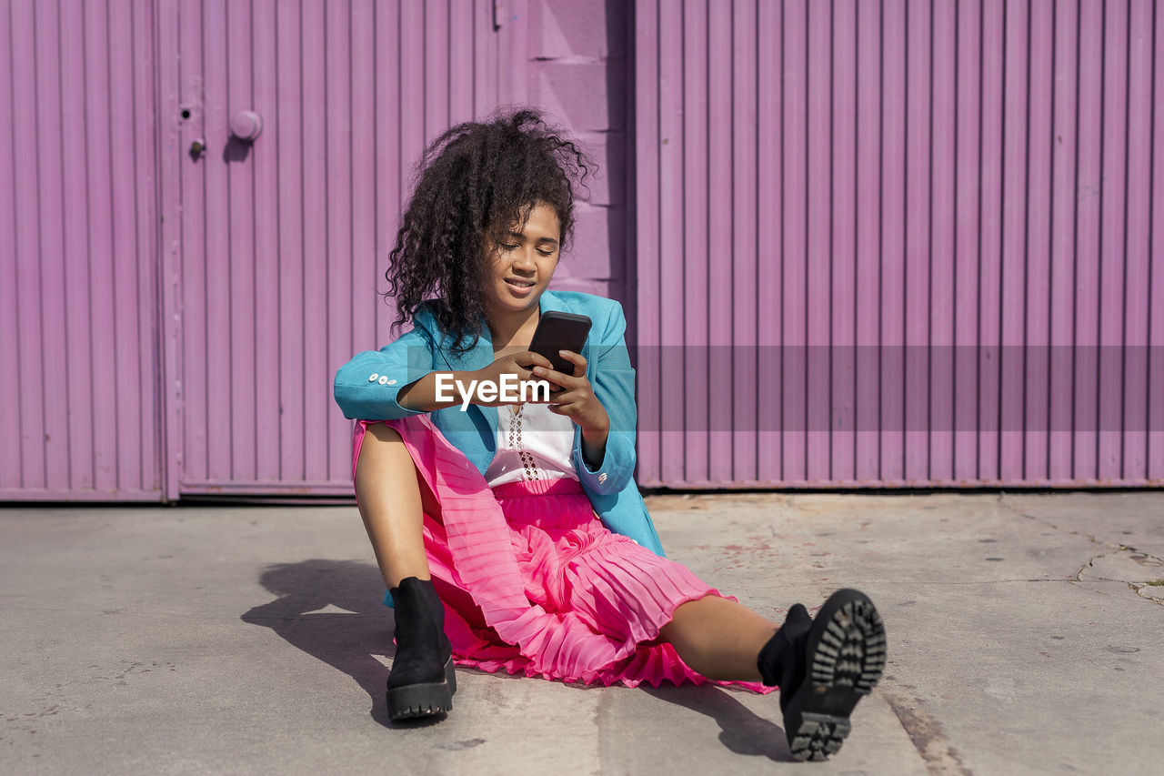 Young woman with afro hairstyle using phone while sitting on footpath