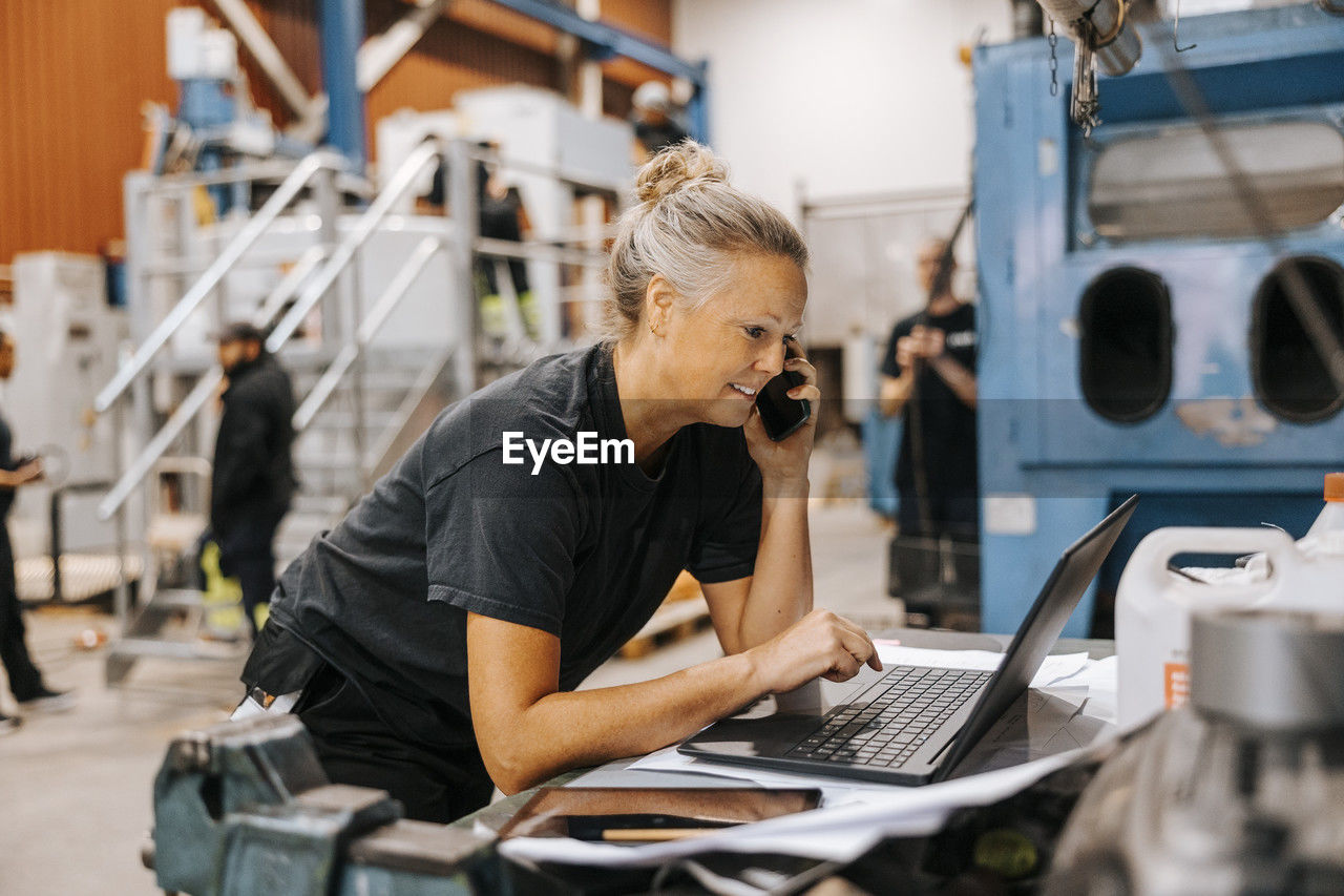 Female warehouse worker talking on smart phone while using laptop in manufacturing factory