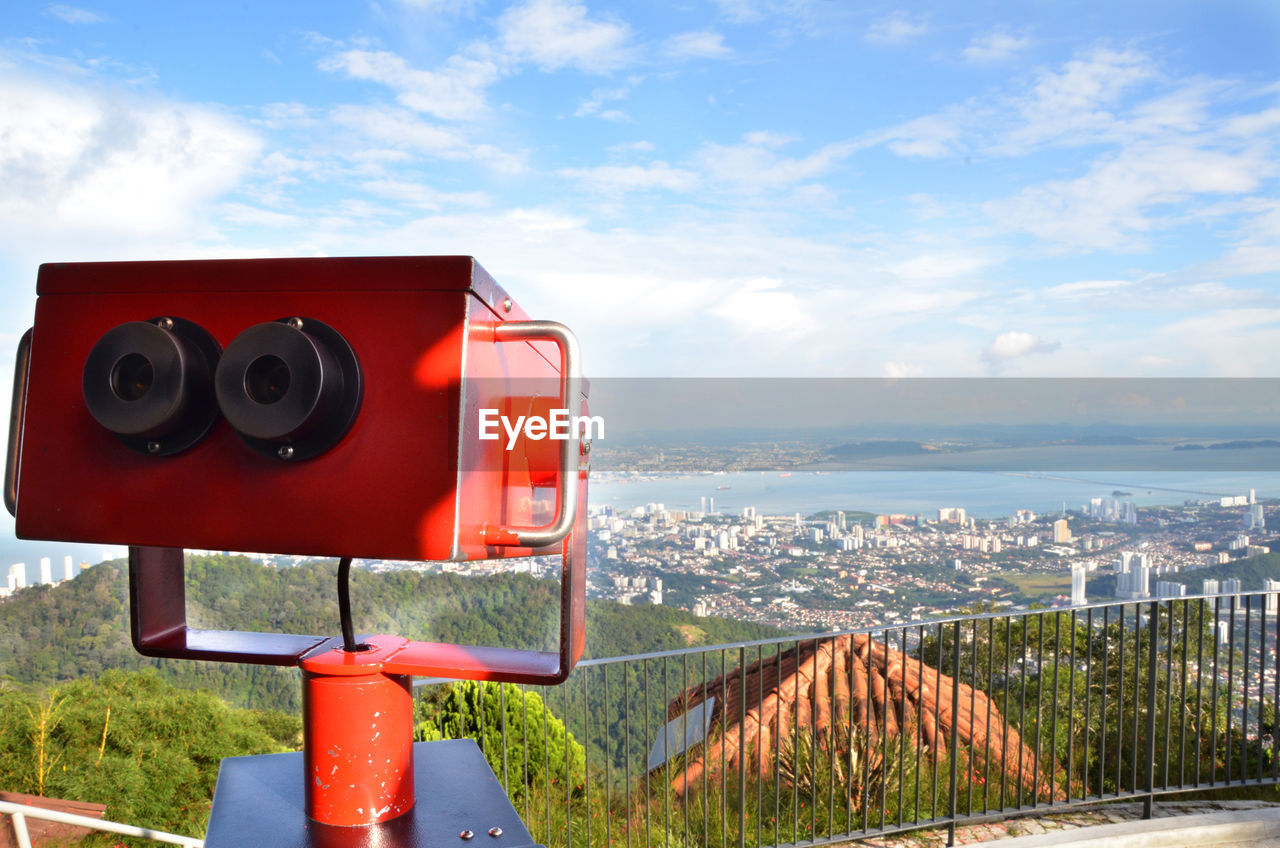 Close-up of red coin-operated binoculars over city against sky