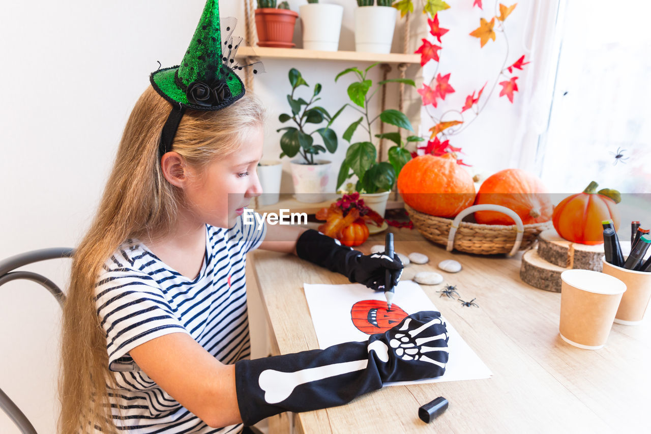 Halloween. a teenage girl in a witch hat drawing a pumpkin sitting by the window