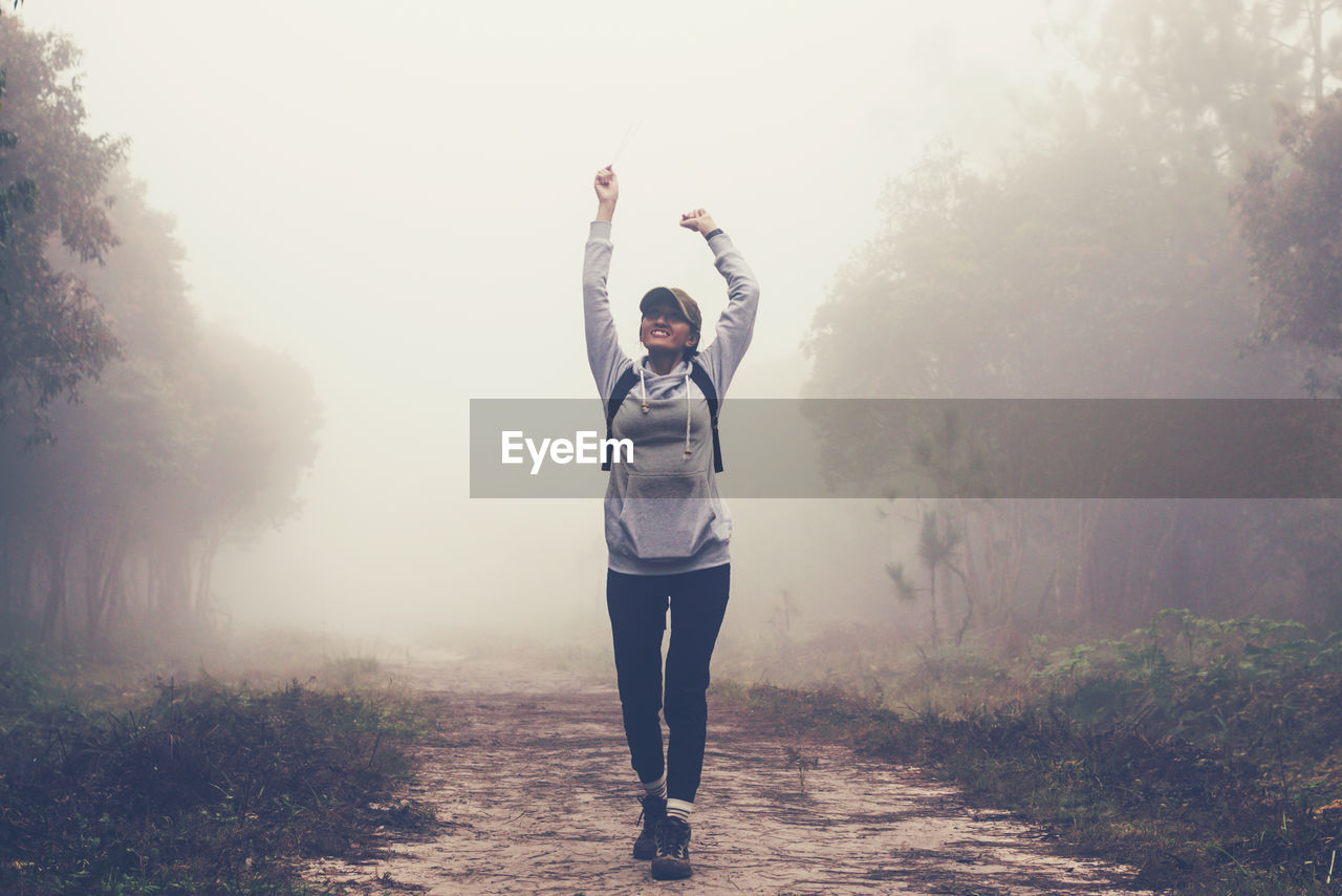 Young woman with arms raised walking in forest during foggy weather