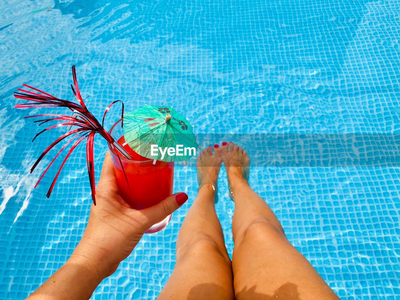 Low section of woman holding drink while sitting by swimming pool