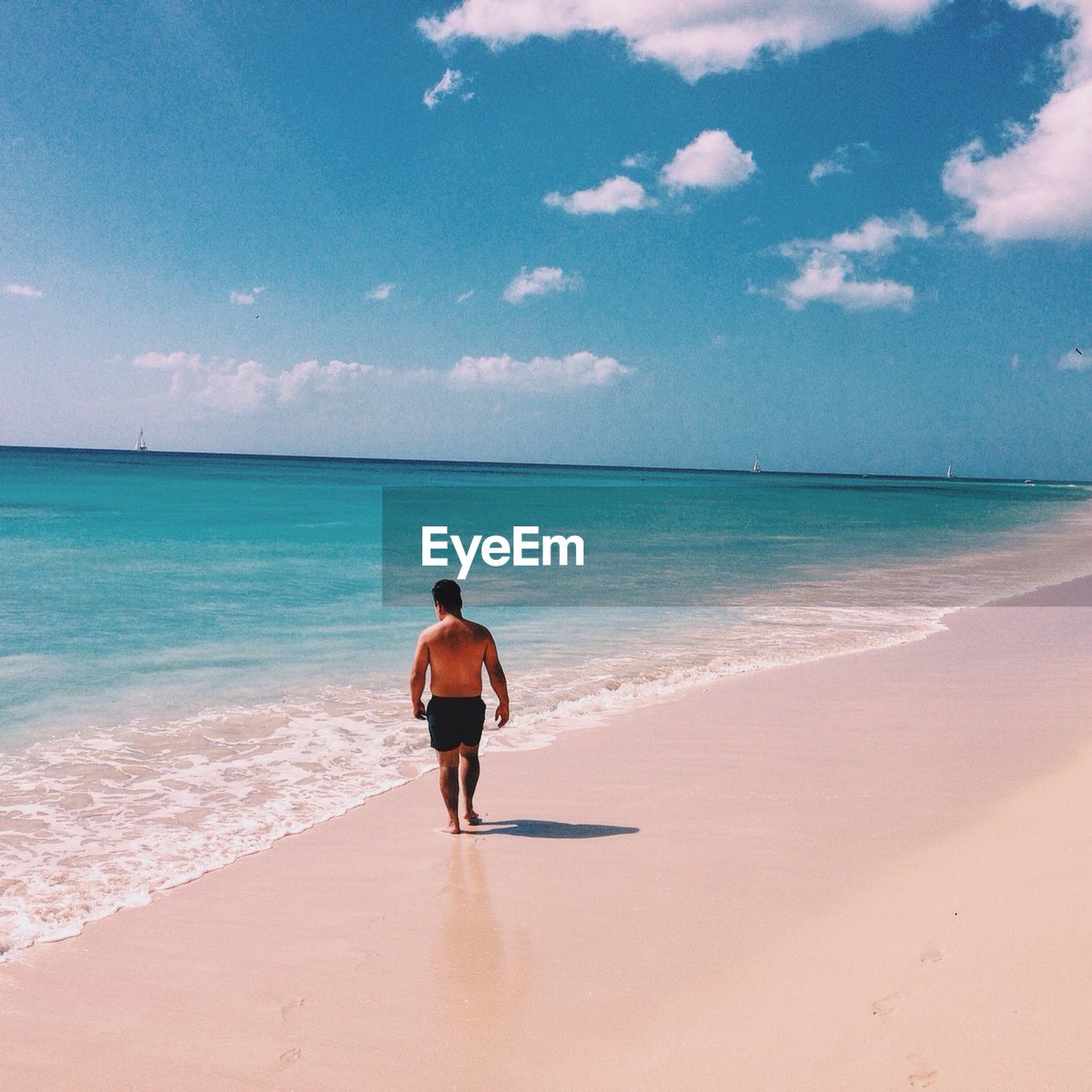 Full length rear view of shirtless man walking at beach against sky on sunny day