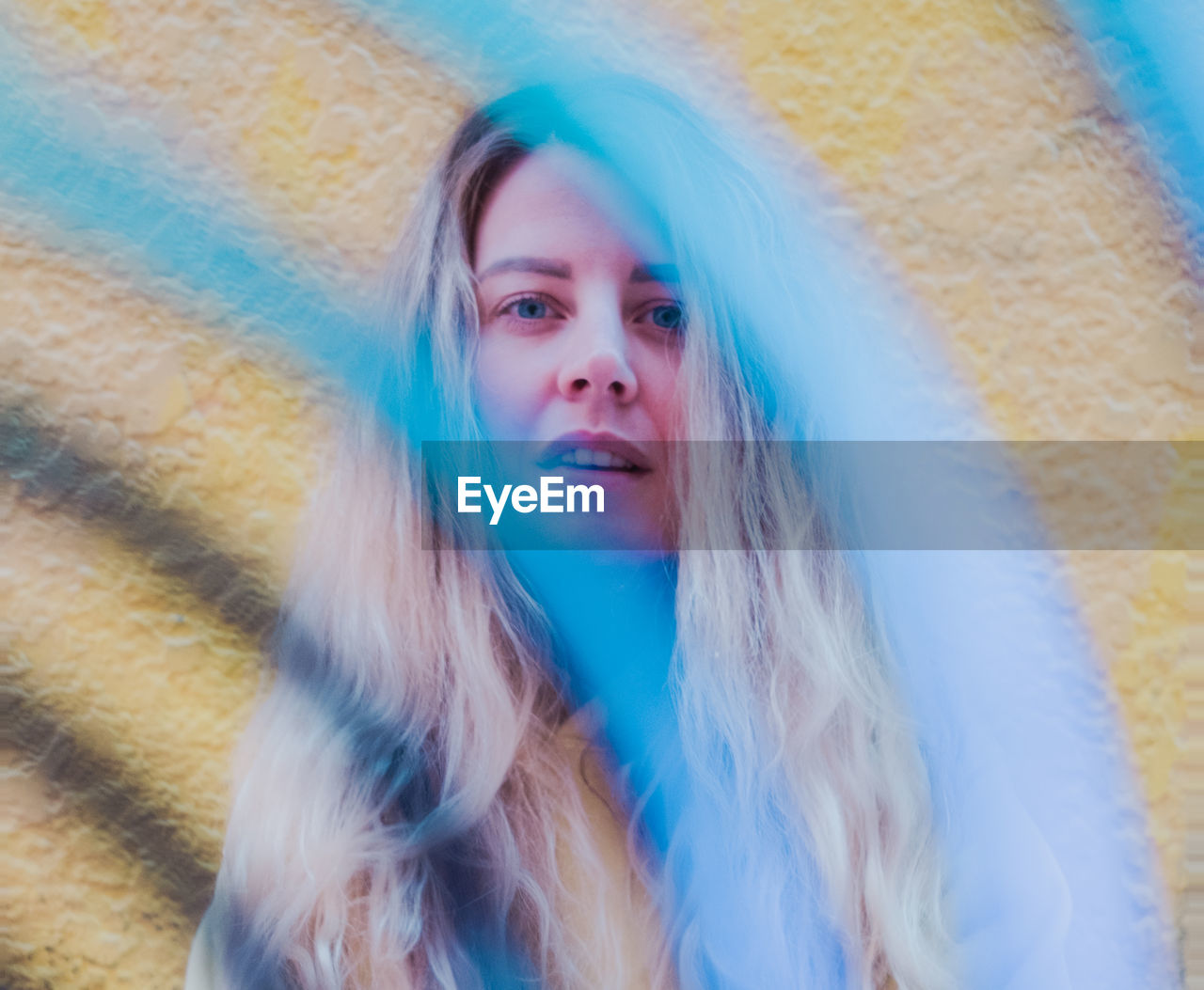 Woman standing against yellow wall. foreground object obscuring image.
blonde hair. blue eyes.