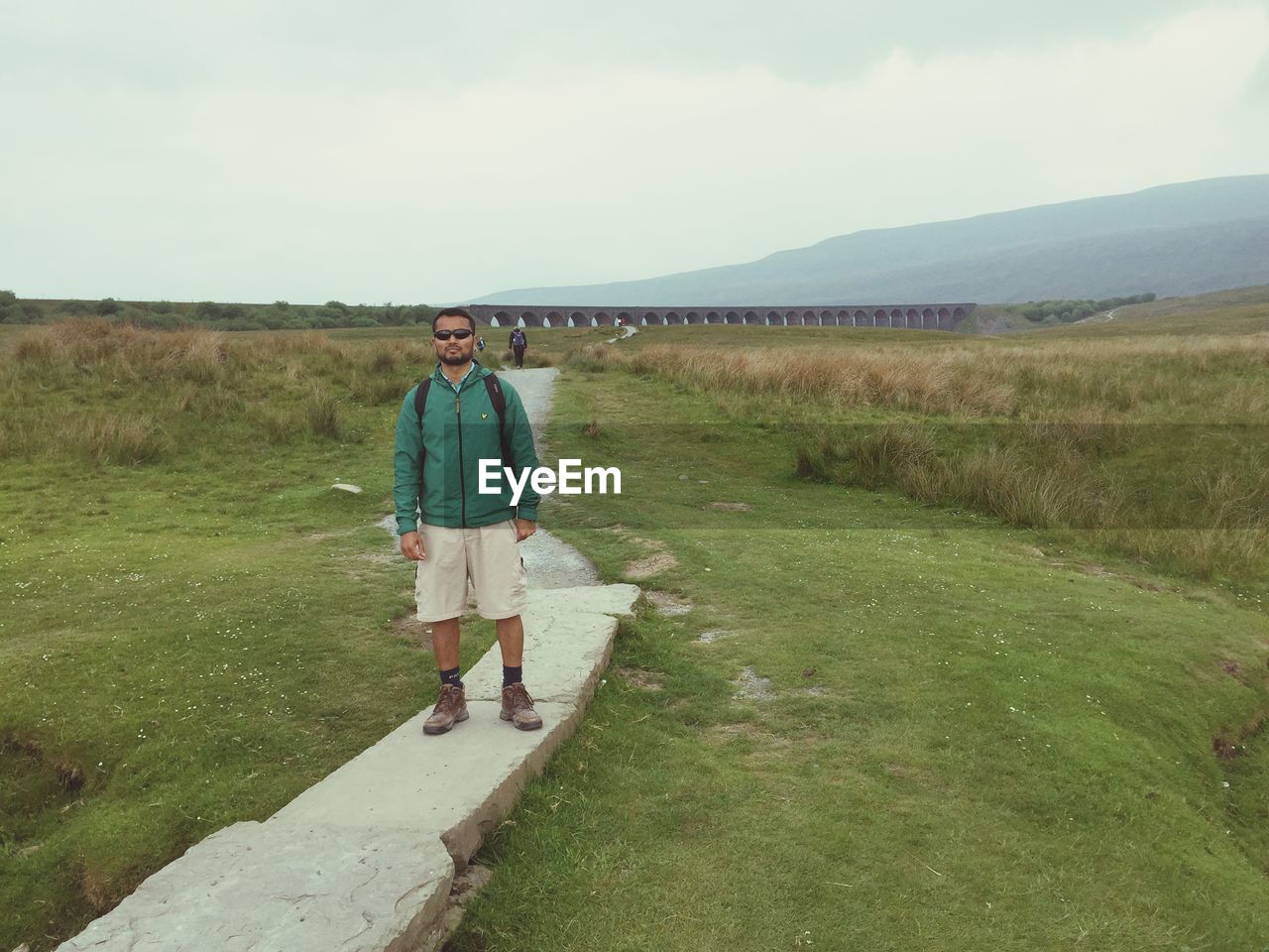 REAR VIEW OF MAN STANDING ON GRASSY FIELD AGAINST SKY