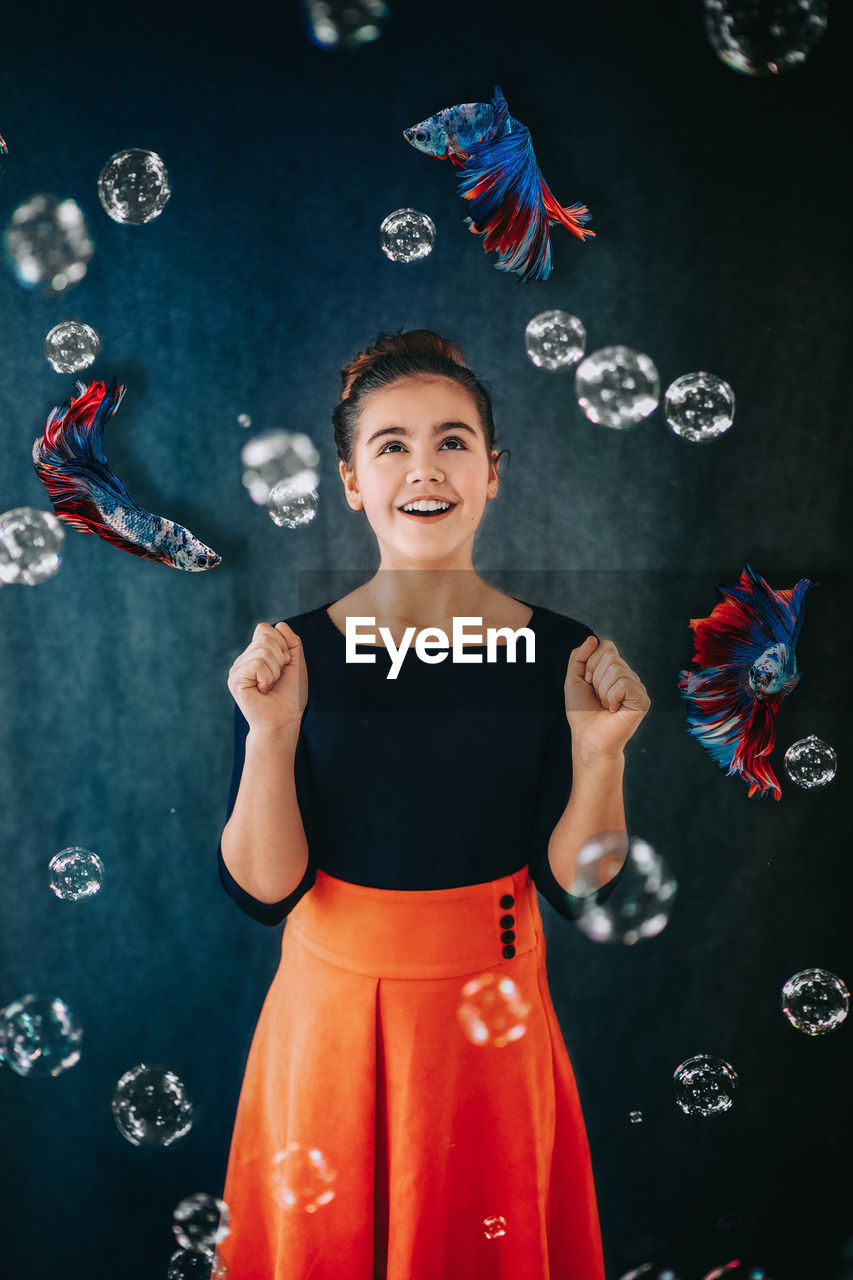 Digital composite image of smiling girl with fishes and bubbles against wall