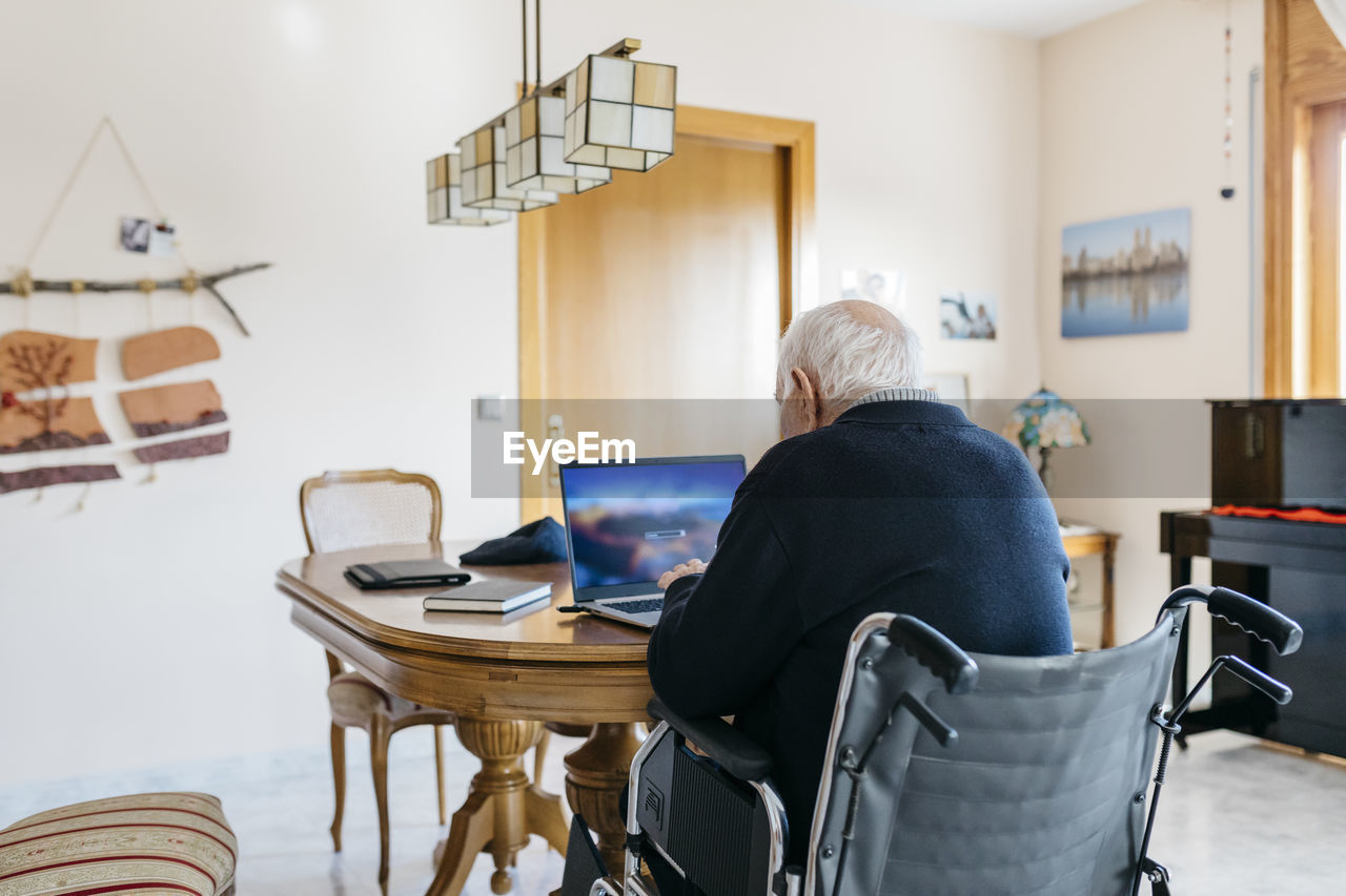 Back view of senior man in wheelchair using laptop at home