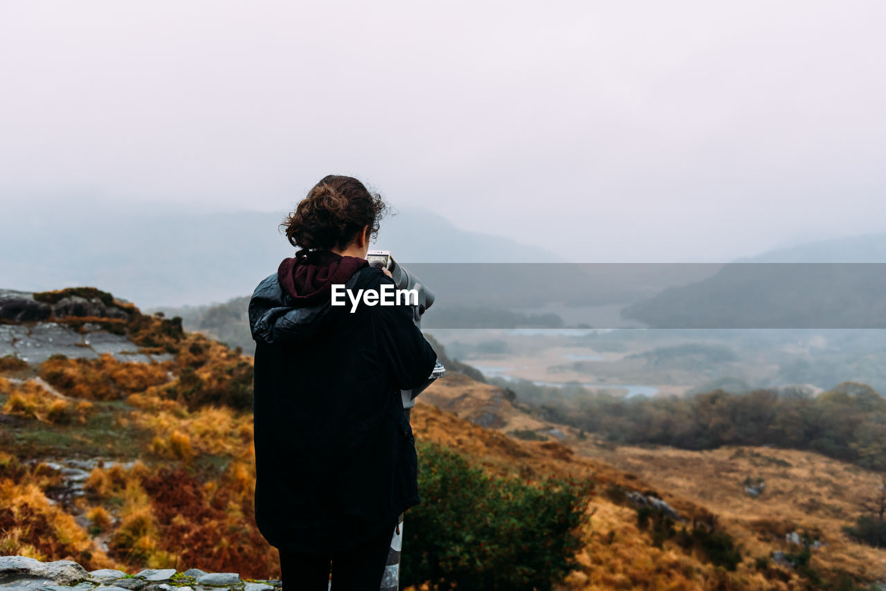 Rear view of teenage girl looking at mountains through telescope against sky