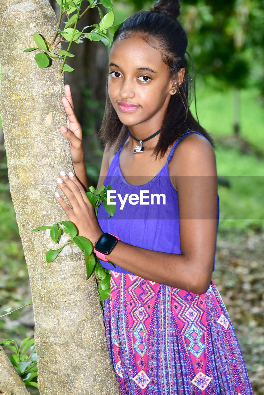 Portrait of young woman standing by tree trunk