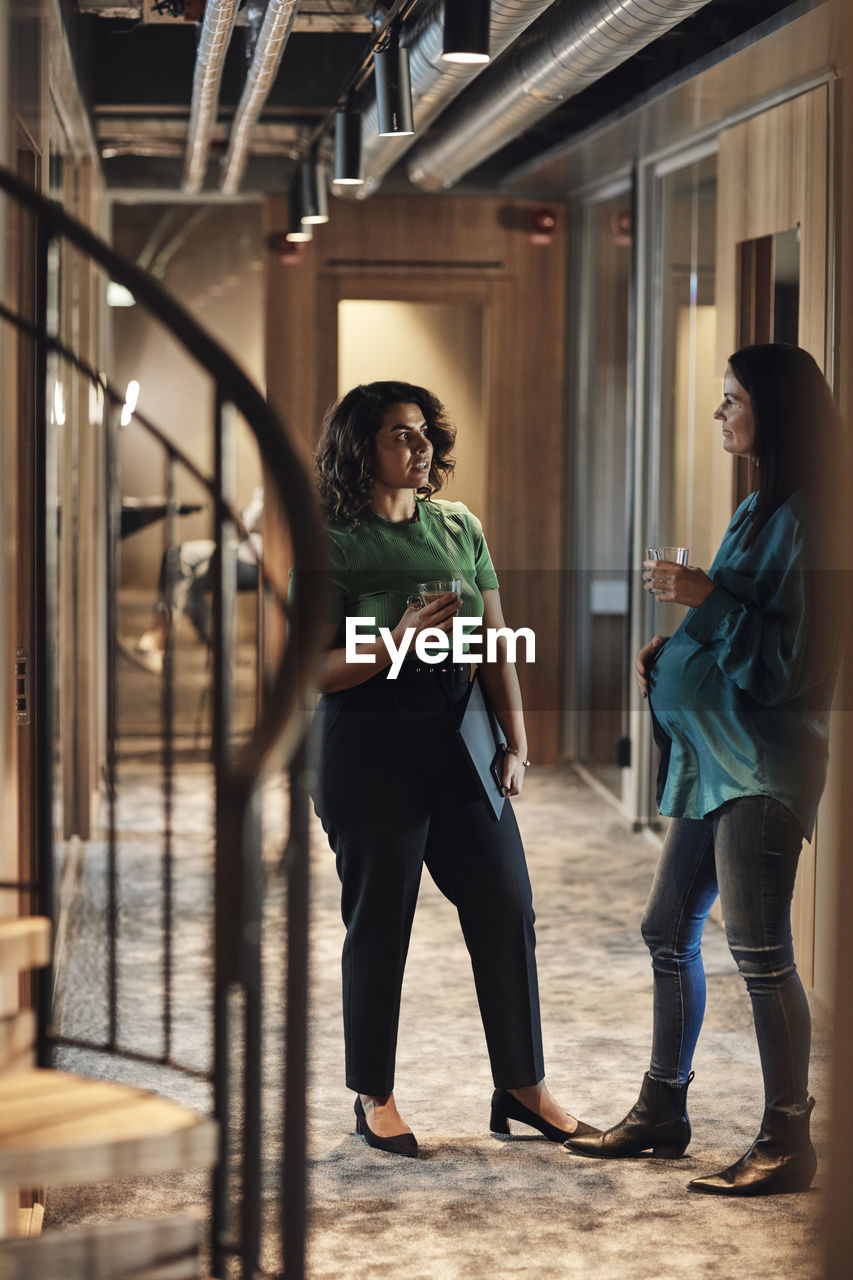 Businesswomen communicating with each other while having drink in corridor at office