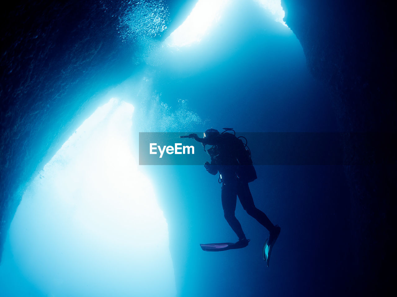 Full body of diver in wetsuit with flashlight in hand swimming underwater of deep sea with clear water near stony formations