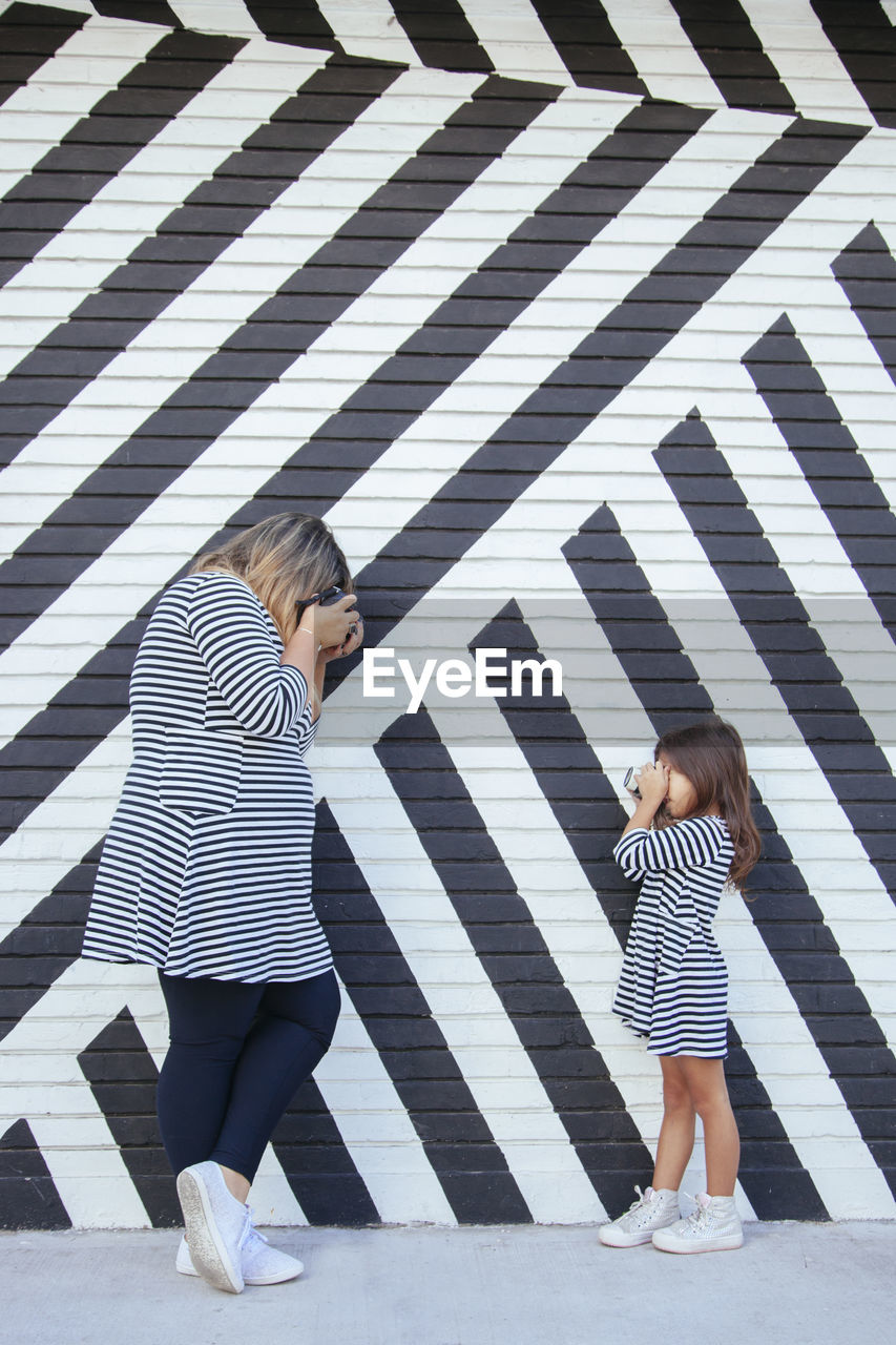 Side view of woman and girl photographing with camera while standing against wall