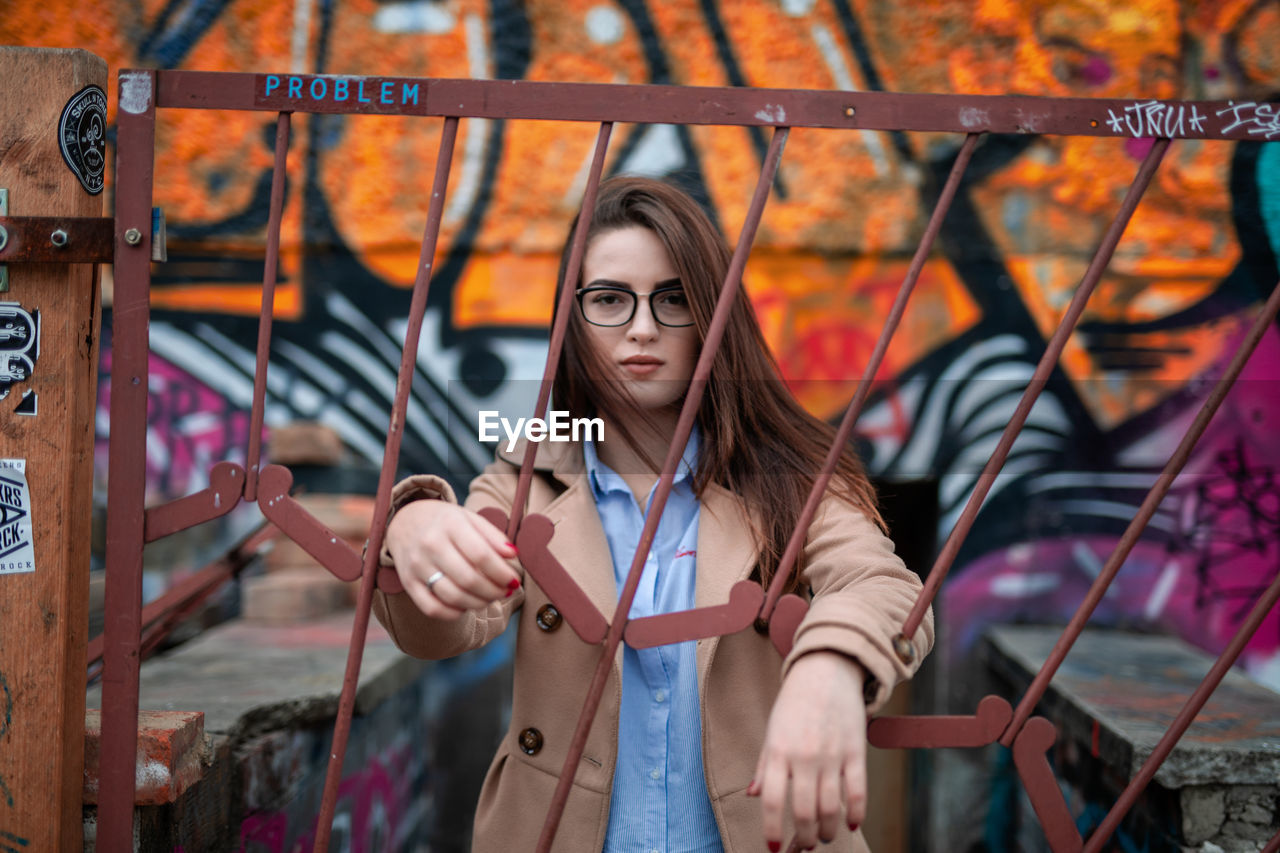 Portrait of young woman standing behind metal gate against graffiti wall