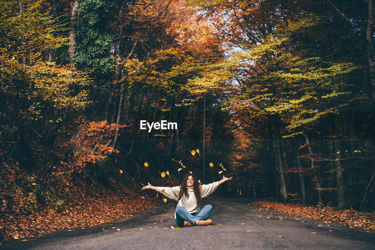 Positive curly haired woman in sweater throwing dry leaves in picturesque autumn forest 