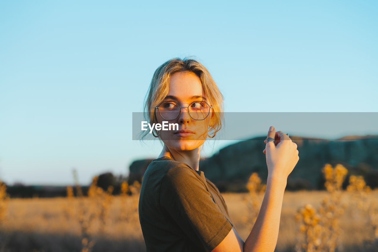 Side view of confident young female traveler in casual clothes and eyeglasses standing in meadow and looking away while relaxing in countryside at sunset