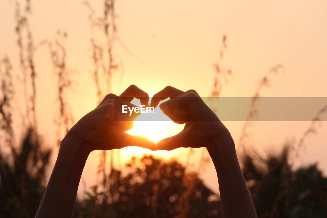 Close-up of hand holding heart shape against sky during sunset