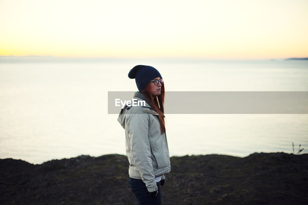 WOMAN STANDING ON BEACH AGAINST SKY DURING SUNSET