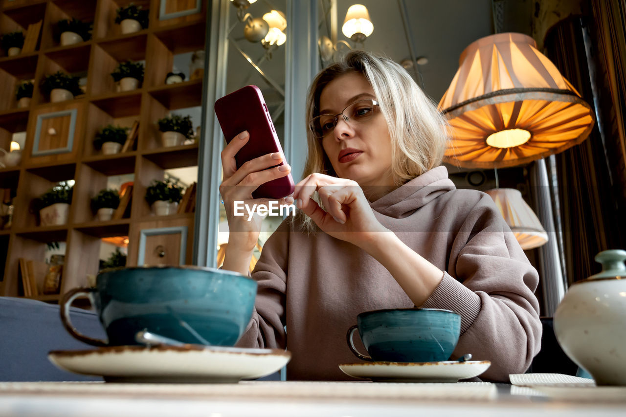 Caucasian middle-aged blonde woman sits in a cafe, drinks coffee and talks on a mobile phone