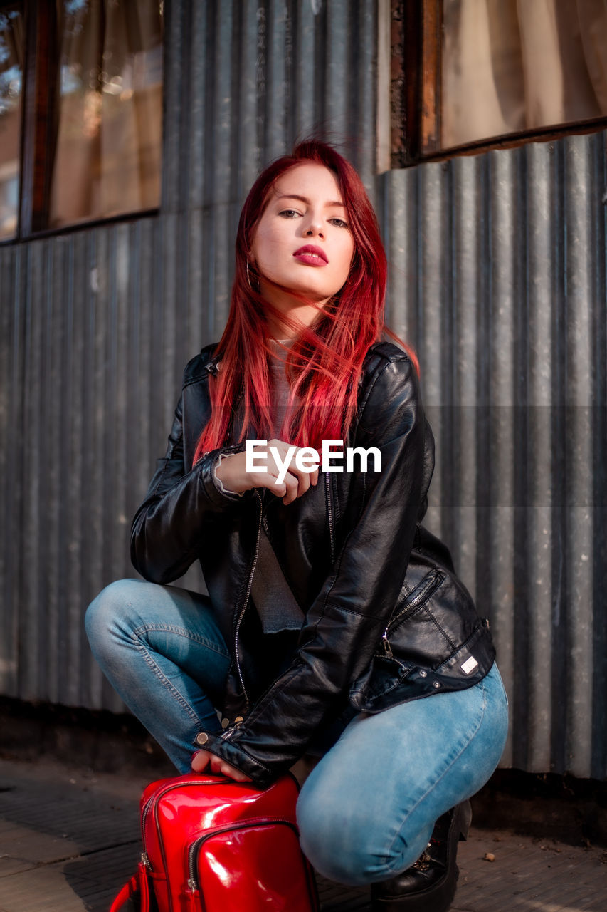 red, one person, women, young adult, adult, clothing, sitting, leather, fashion, portrait, leather jacket, female, architecture, city, lifestyles, jacket, casual clothing, hairstyle, person, long hair, city life, redhead, looking at camera, leisure activity, full length, emotion, relaxation, photo shoot, luggage and bags, front view, footwear, dyed red hair, human face, arts culture and entertainment, street, looking, jeans, brown hair, outdoors