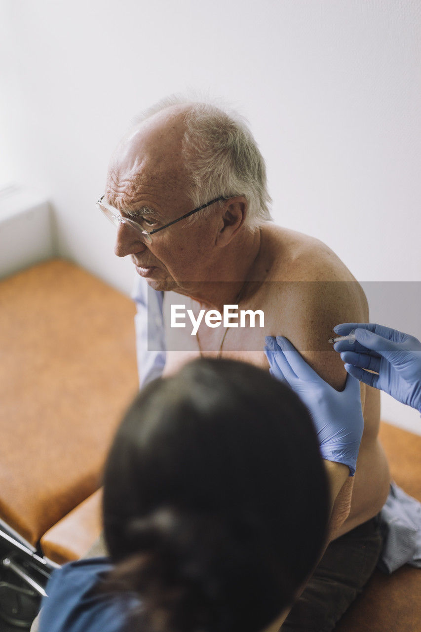 Shirtless male senior patient taking vaccination on arm at hospital