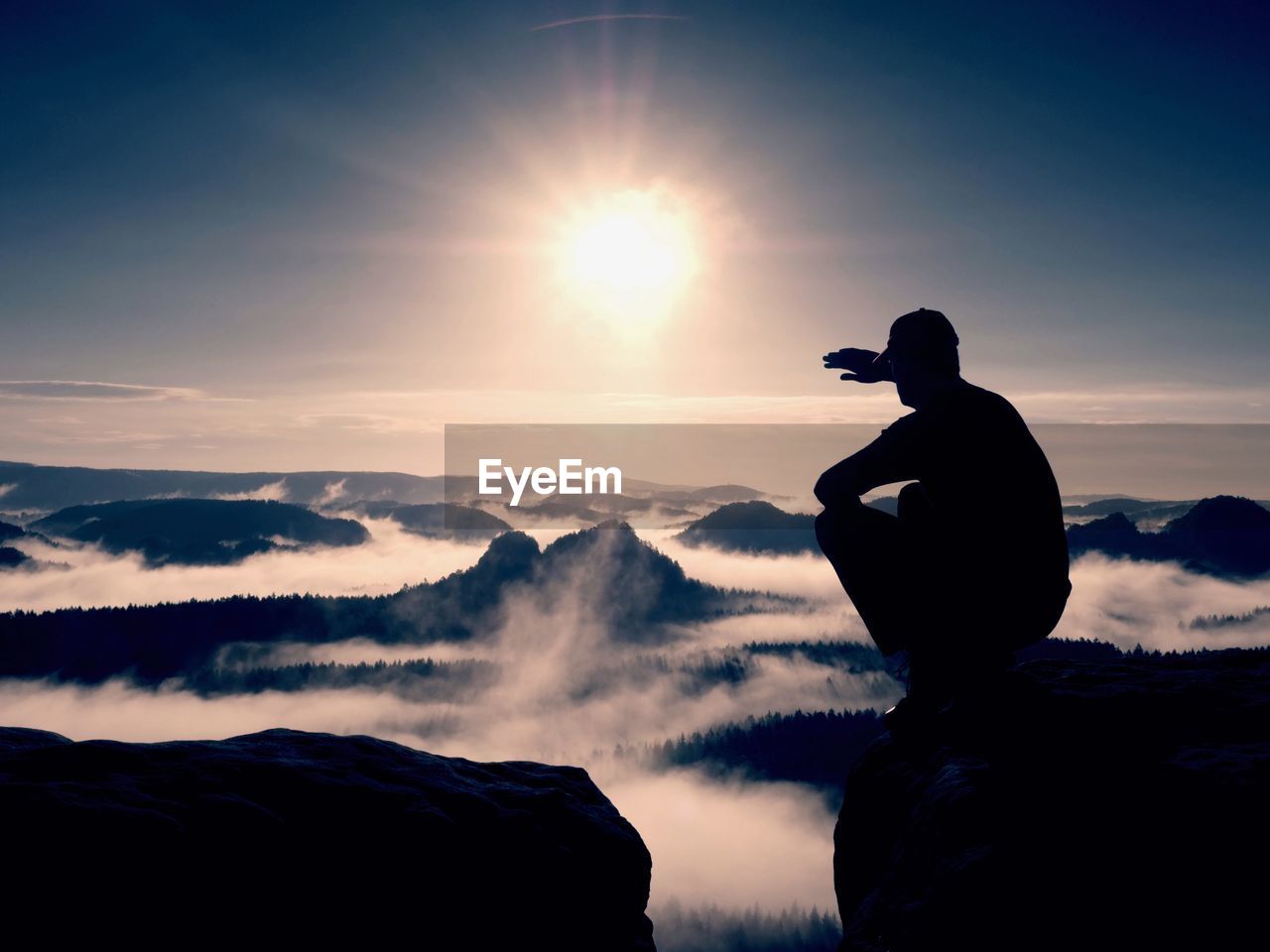 Silhouette of alone hiker in black and cap sit on cliff. blue filter photo. man looking to mist 