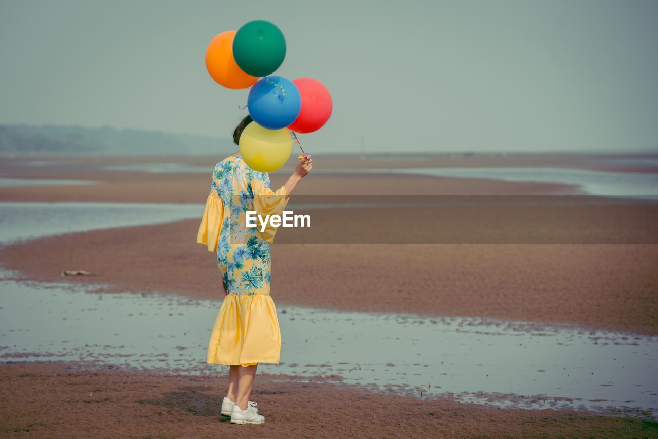 Rear view full length of young woman holding colorful balloons at beach