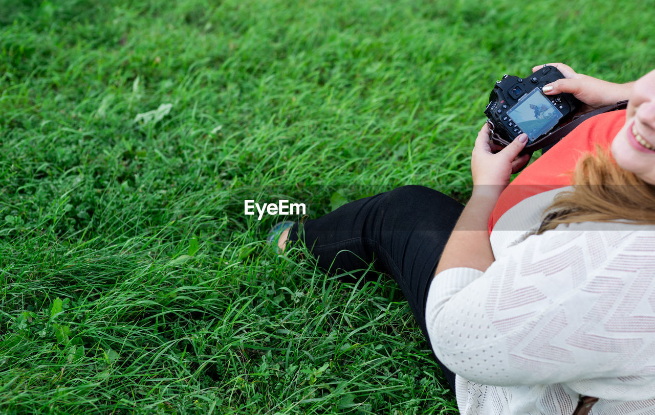Low section of woman holding camera while sitting on grass outdoors