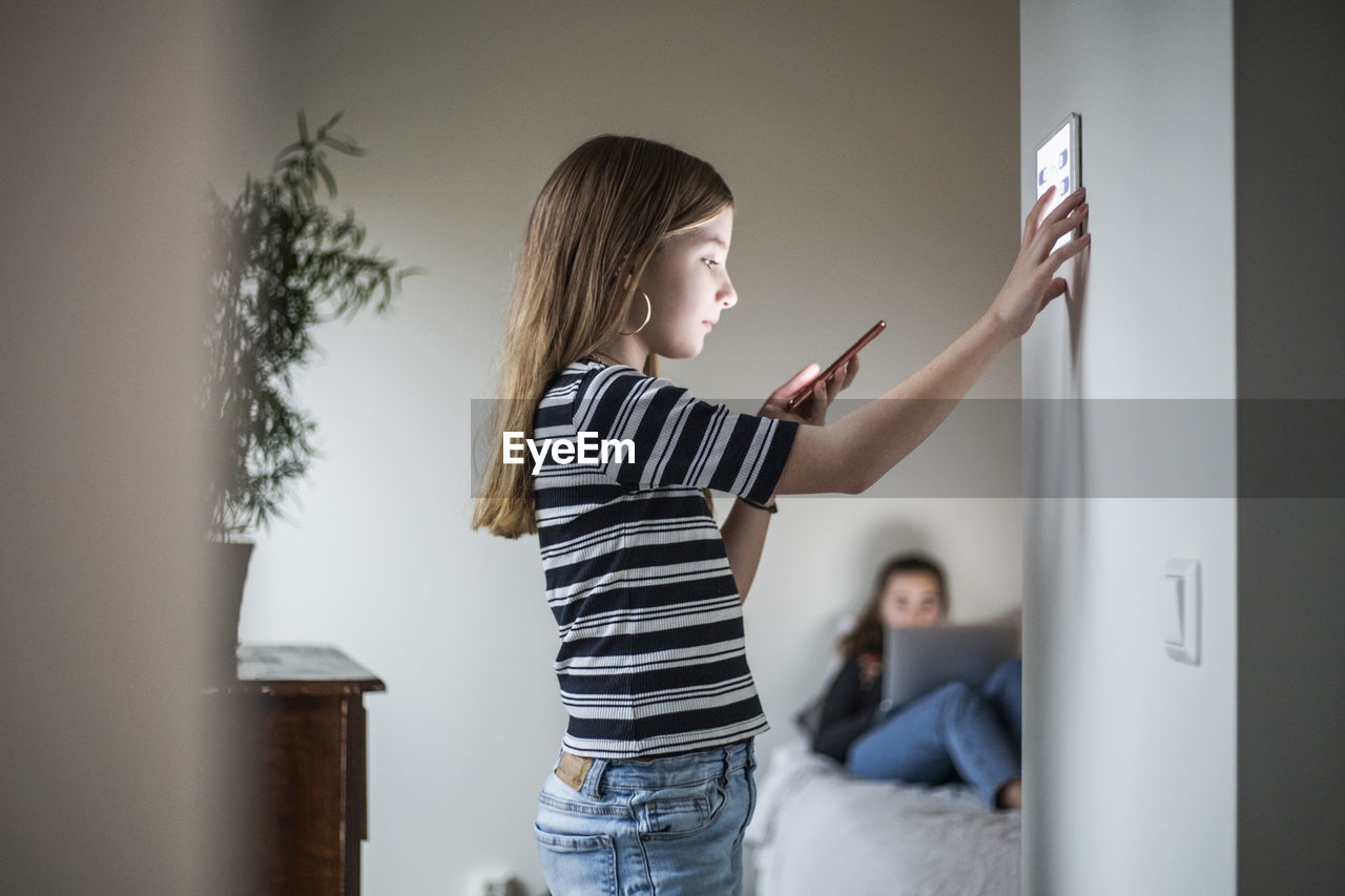Girl looking at smart phone while using digital tablet on wall with sister in background at smart home