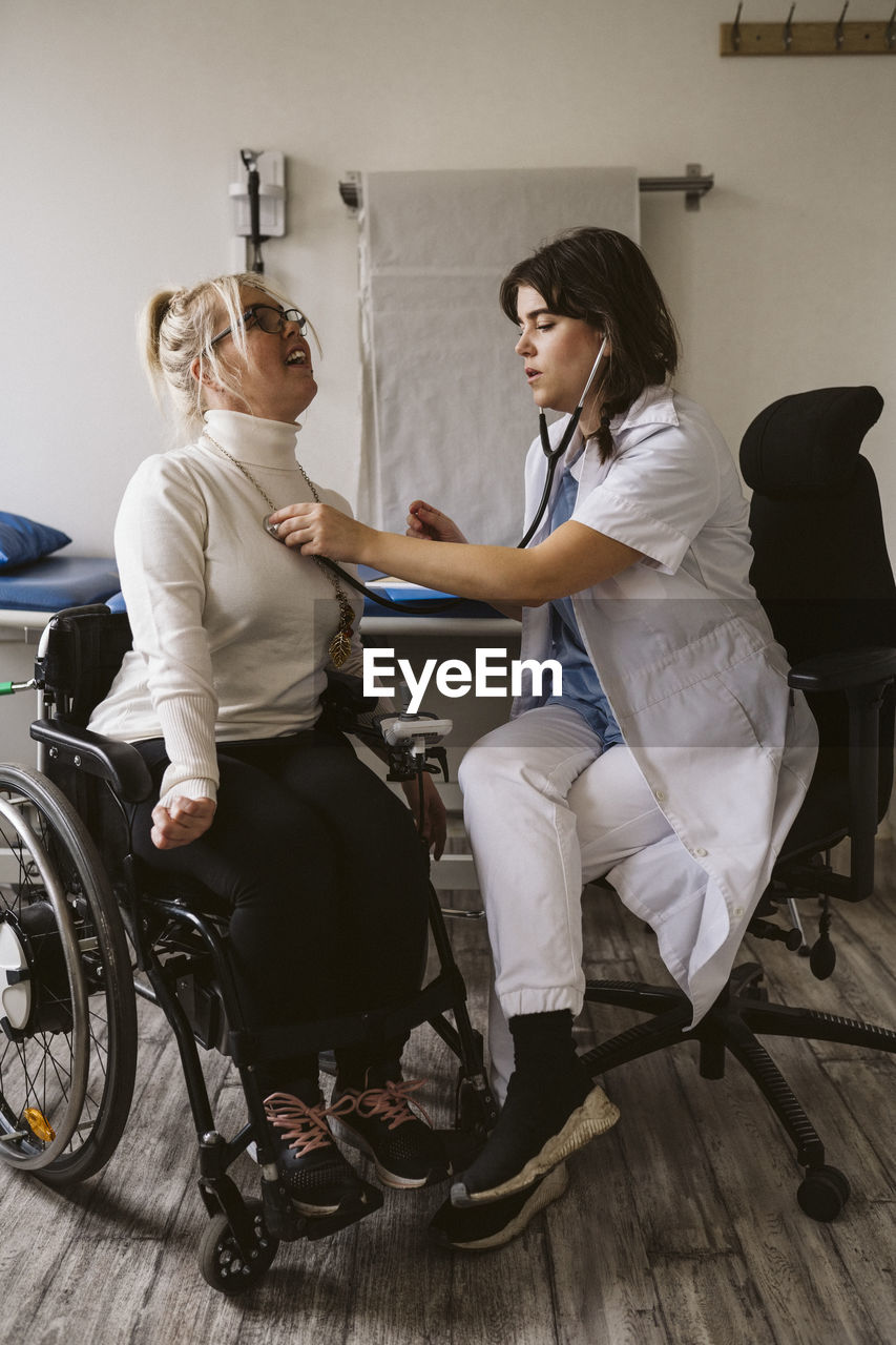 Full length of female healthcare worker examining disabled patient with stethoscope in medical clinic