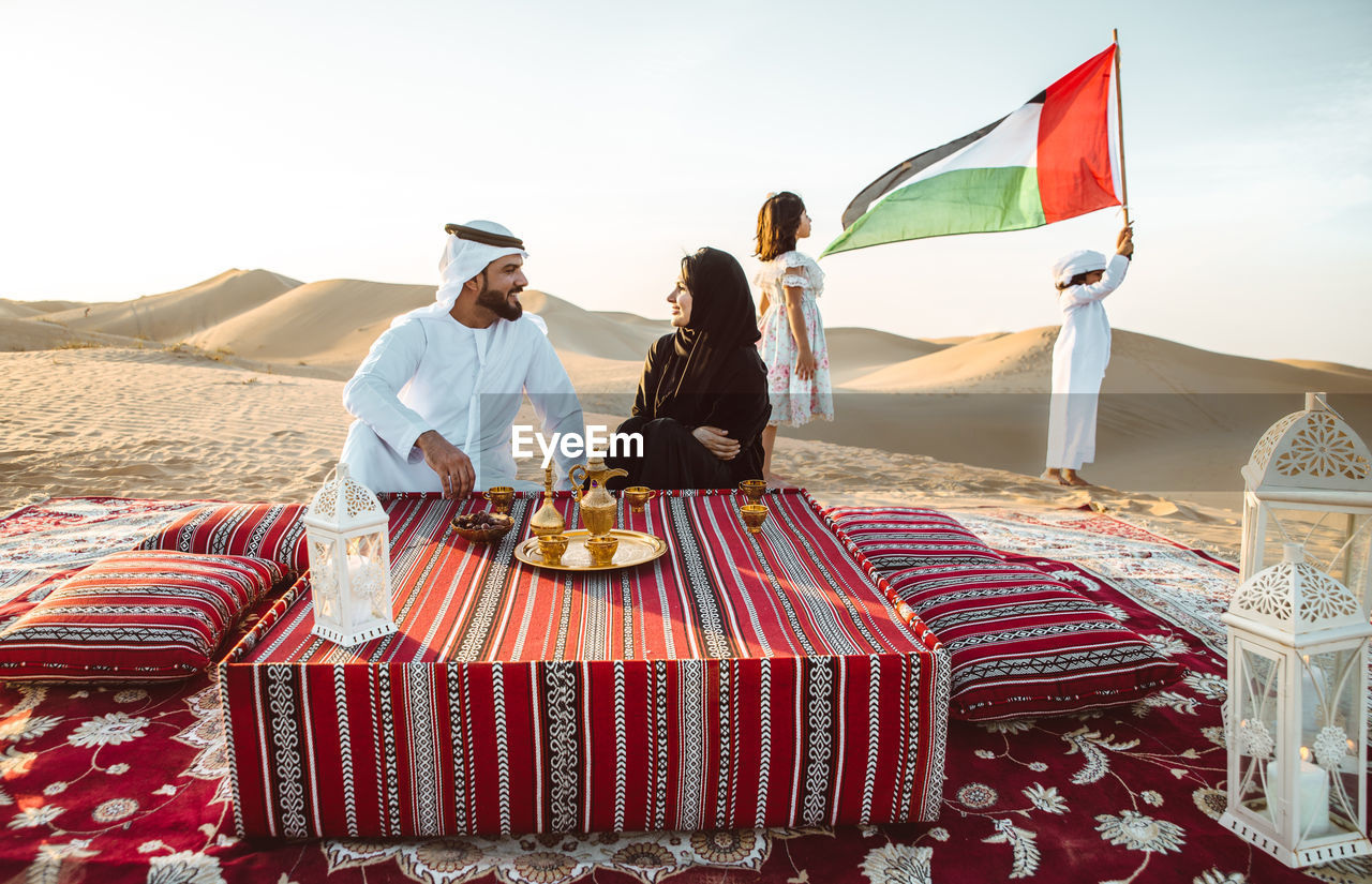 Happy parents with tea on carpet against children with united arab emirate flag sat desert