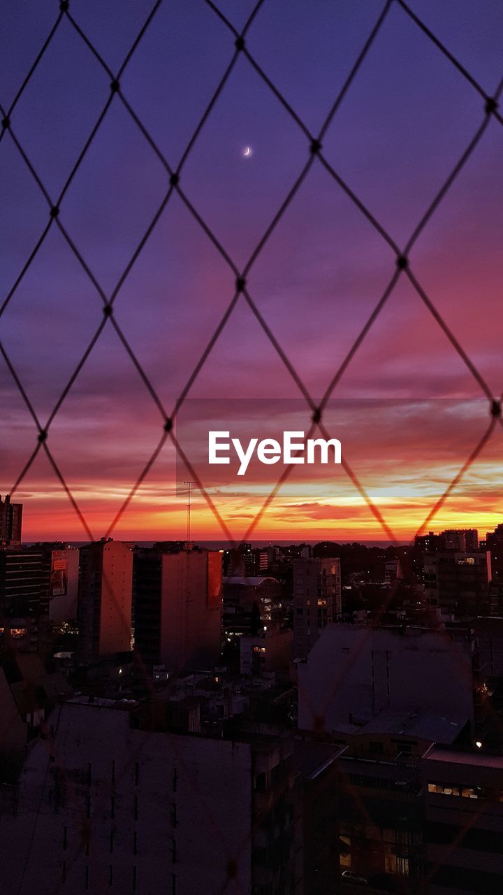 Cityscape seen through chainlink fence against sky at sunset