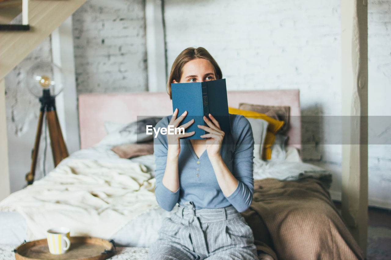 FULL LENGTH OF WOMAN SITTING ON BED IN BEDROOM