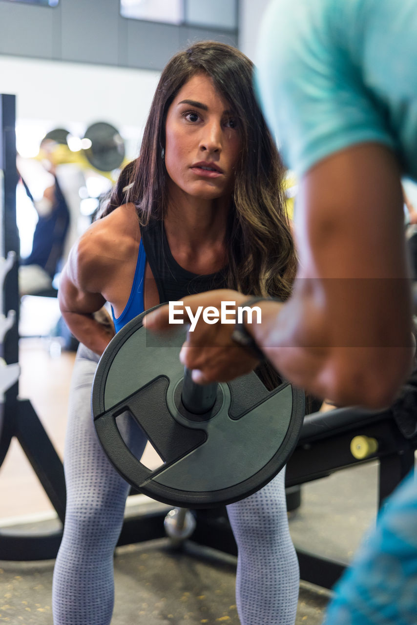 Concentrated young ethnic female in sportswear doing exercises with barbell with support of personal instructor during training in gym