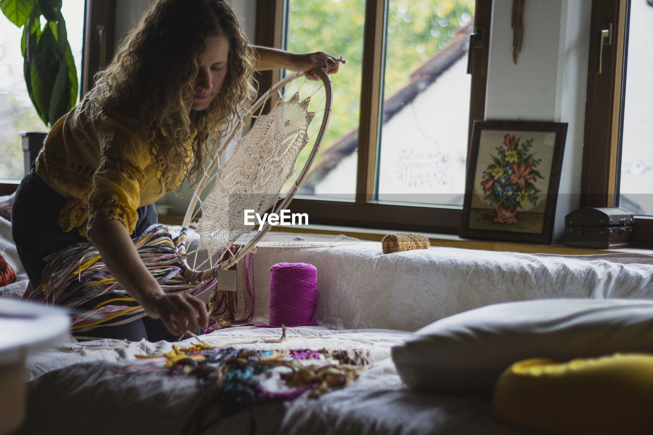 Side view of woman creating handmade dreamcatcher with long threads spending time in house