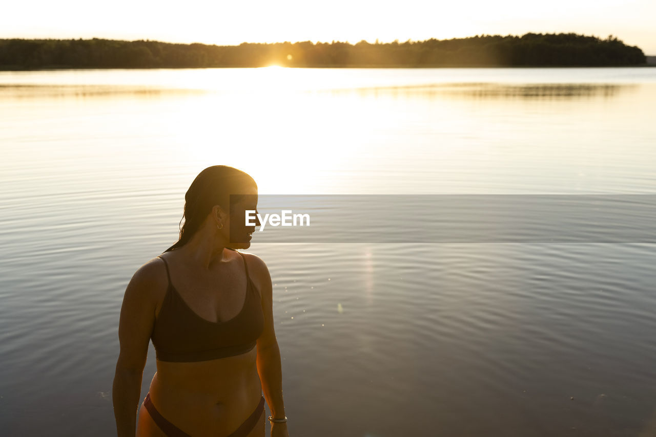 Woman in bikini looking away while standing against lake during vacation