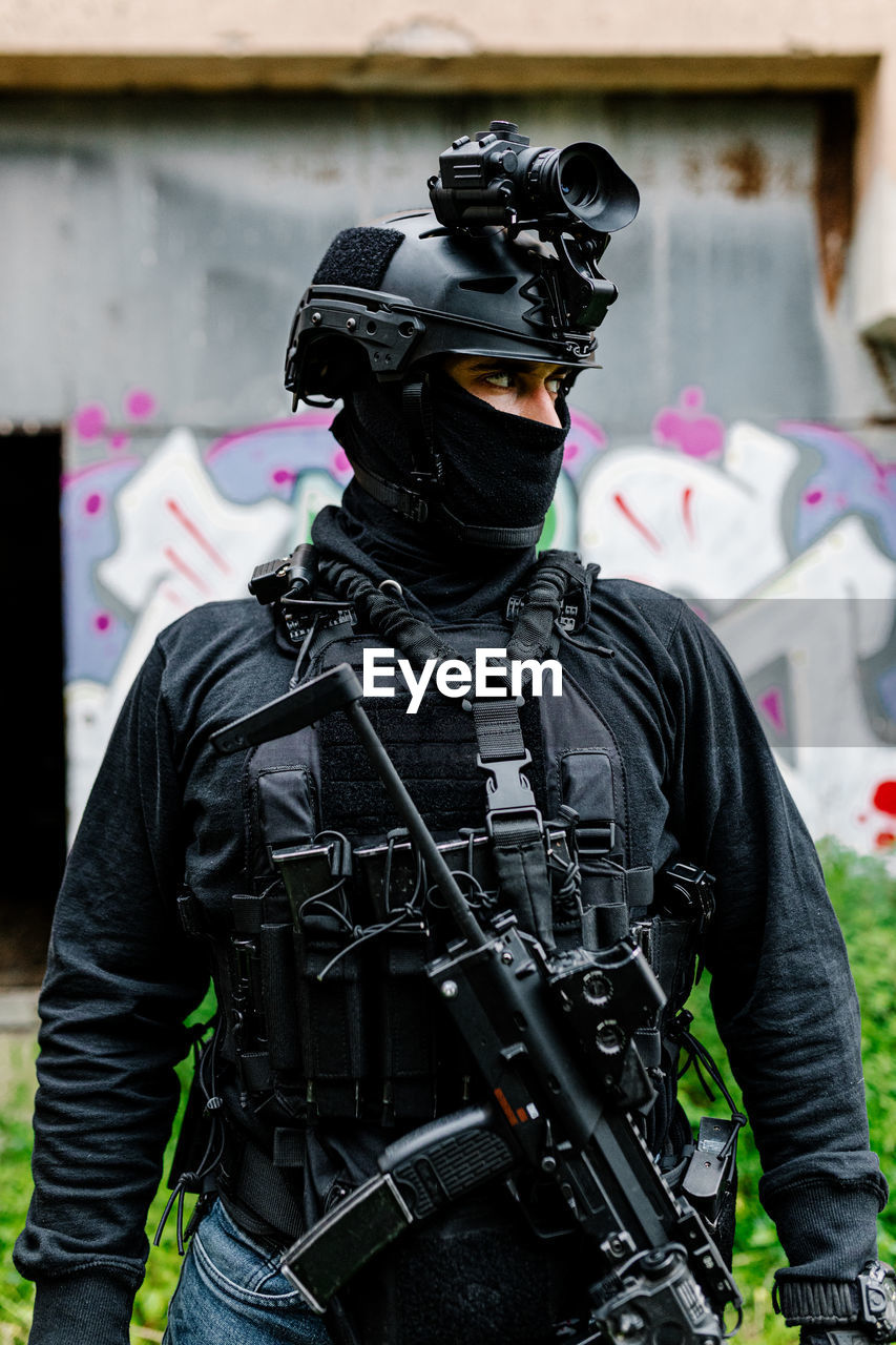 Serious young unrecognizable male warrior in modern protective wear balaclava and helmet with professional rifle looking away while standing near old building with graffiti on metal wall