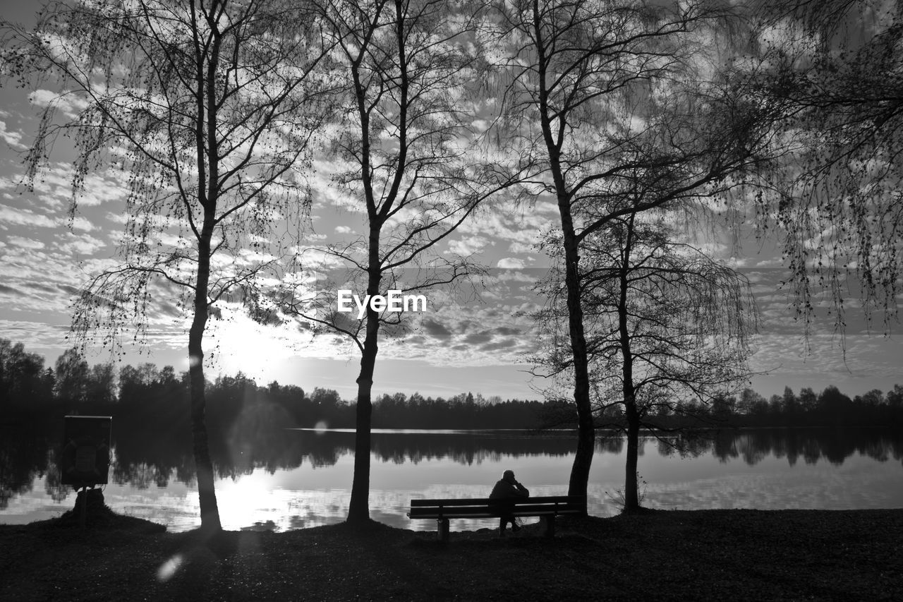 Silhouette man sitting on bench at lakeshore against sky