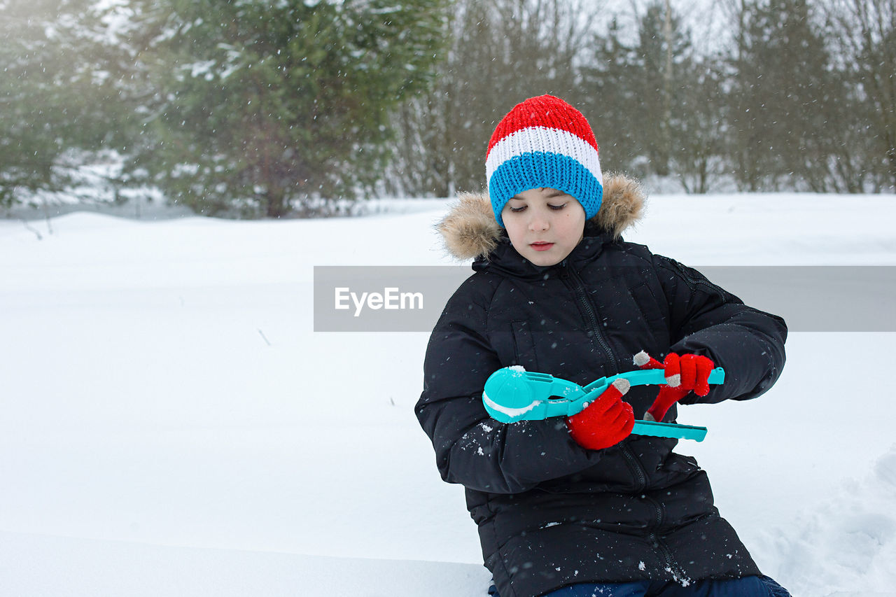 A boy sits in the snow, makes snowballs using a light blue plastic sculpting tool