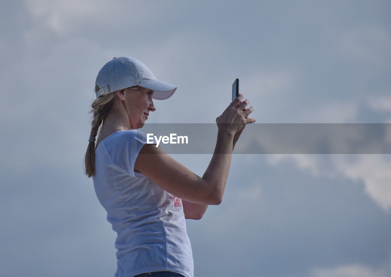 Side view of woman photographing while standing against cloudy sky