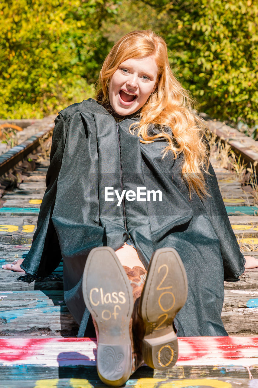Portrait of smiling girl in graduation gown sitting on wood with text and numbers on shoes