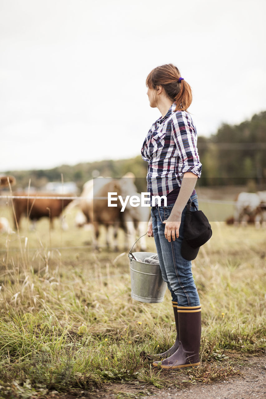 Female farmer with bucket standing on field with animals grazing in background